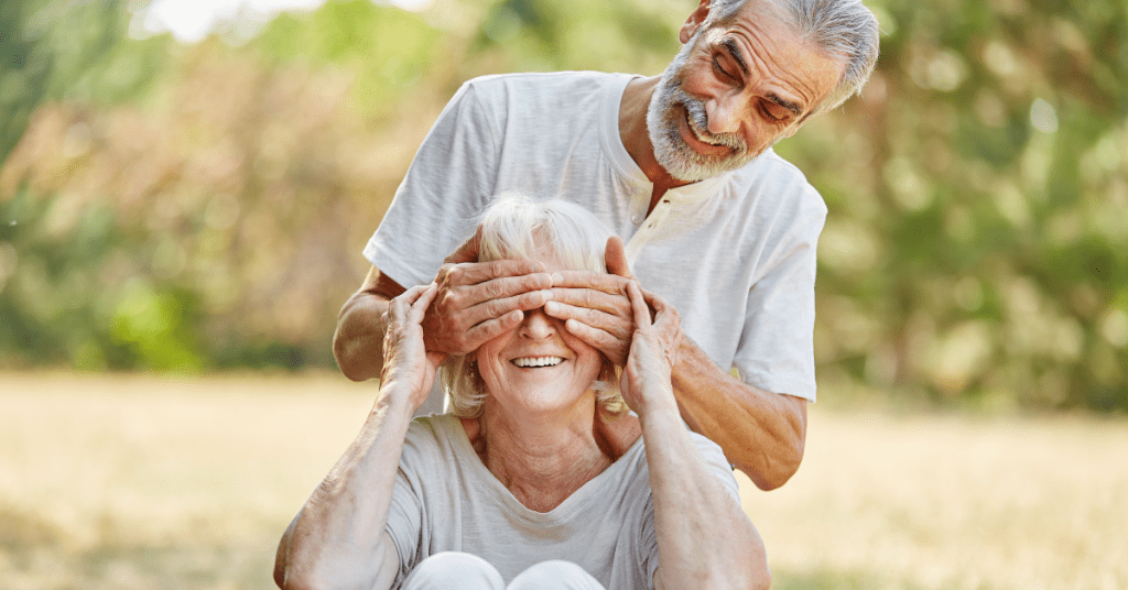 older man covers eyes of older woman in field while quizzing her on eye health